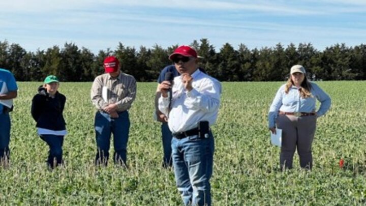 Speaker addressing a group in a green field under a clear blue sky.